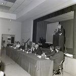 Man Speaking on Stage above Panel Table for Master Charge by George Skip Gandy IV