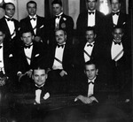 Bulletin board photo of photo of posed group of men in tuxedos and sashes; three rows of men, tall windows, wood floor-possibly taken in a ballroom