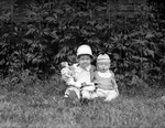 Betty and Dick wearing hats and sitting outside in grass in front of leaf wall. Betty holds doll by Francis G. Wagner and Nelson Poynter Memorial Library