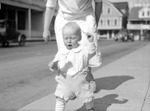 Baby Bud toddling on Maine sidewalk away from Irma; street, cars, buildings in background. Date: 1927