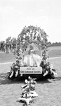 Girl sitting on a flower-covered float with sign, "Just an Old Fashioned Garden." People standing in the background; Envelope said photo was taken in Sunken Gardens by Francis G. Wagner and Nelson Poynter Memorial Library