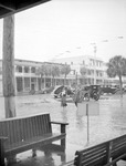 Man and woman with umbrella crossing brick street (Central Avenue), possibly in the rain; Benches in foreground, cars and unnamed buildings in background by Francis G. Wagner and Nelson Poynter Memorial Library
