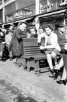 People sitting on green benches in front of Rexall Drugs by Francis G. Wagner and Nelson Poynter Memorial Library
