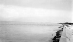 Seawall with two people walking on it; water with waves and birds; sandy soil with grasses by Francis G. Wagner and Nelson Poynter Memorial Library