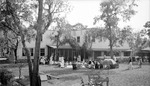 Large group of children and women in yard behind large two-story building with glass lanai, possibly a school, many sitting in chairs; flowered umbrella standing, wooden dollhouses under trees, seesaw by Francis G. Wagner and Nelson Poynter Memorial Library
