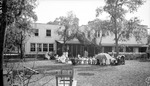 Large group of children and women in yard behind large two-story building with glass lanai, possibly a school, many sitting in chairs; lowered umbrella standing. wooden dollhouses under trees, seesaw by Francis G. Wagner and Nelson Poynter Memorial Library