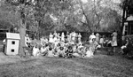 Large group of children and women in yard (two-story building, possibly a school, barely visible), some sitting in chairs; wooden dollhouse under tree, cars in background by Francis G. Wagner and Nelson Poynter Memorial Library