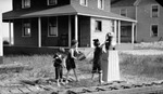 Three unknown barefoot children with pails at a water pump next to railroad tracks. One girl is pumping water into pail while the second girl splashes her; houses clearly visible in background by Francis G. Wagner and Nelson Poynter Memorial Library